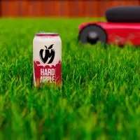 A 3D rendered scene featuring a beverage can, realistic grass, lawnmower, and fence in the distance.